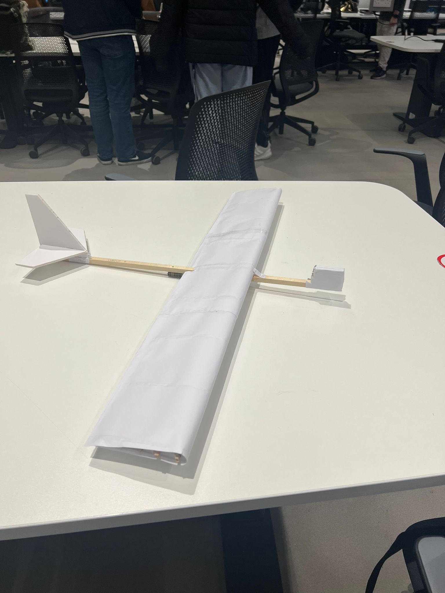 White model aircraft with a long wooden fuselage, wing and tail on a university workbench.